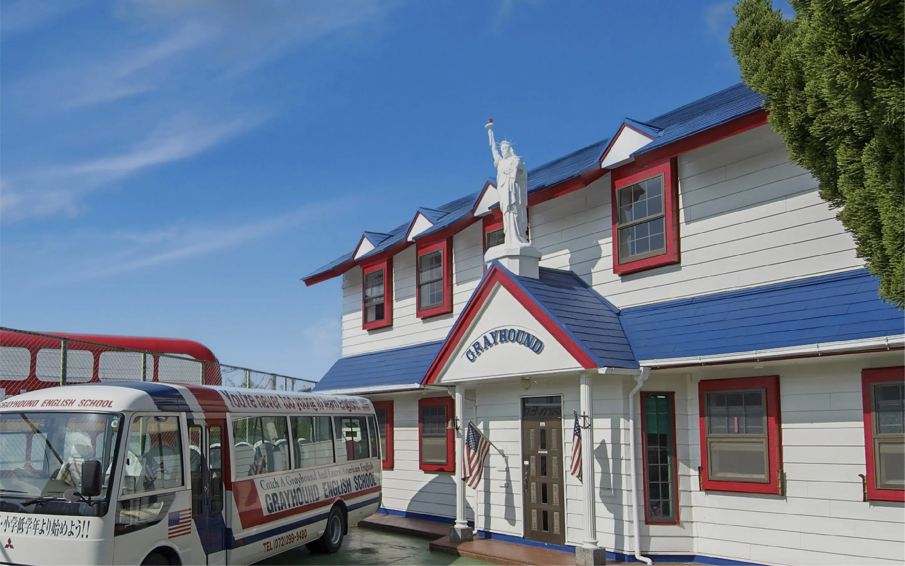 青空の下、自由の女神像が屋上にそびえる赤い縁と青い屋根の校舎とグレイハウンドイングリッシュスクールの送迎バス